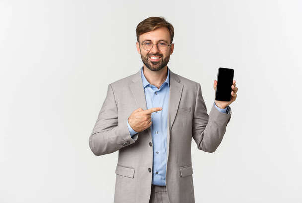 Красивый улыбающийся бизнесмен с бородой, в сером костюме и очках, указывающий пальцем на экран мобильного телефона, показывающий приложение, стоящий на белом фоне - Фото, изображение