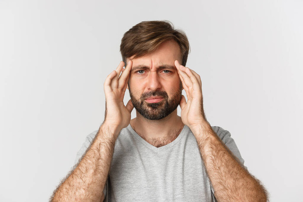 Lähikuva synkkä parrakas mies, koskettava pää ja irvistys, jolla on päänsärky tai migreeni, seisoo valkoisella taustalla - Valokuva, kuva