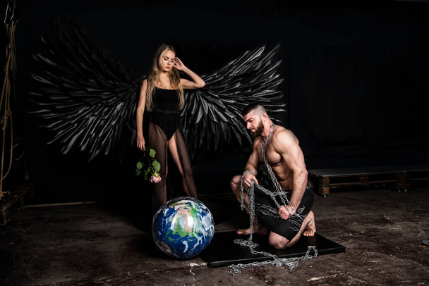 beautiful girl in angel costume with black wings and handsome muscular man struggling with difficulties on a black background - Photo, Image