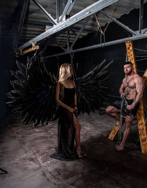 krásná dívka v andělském kostýmu s černými křídly a hezký svalnatý muž zápasí s obtížemi na černém pozadí - Fotografie, Obrázek
