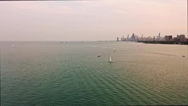 Panorama aéreo en vista de una lancha a motor que cruza un tranquilo lago Michigan mientras los veleros flotan antes del atardecer con el horizonte de Chicago y los edificios de gran altura en el horizonte más allá. - Imágenes, Vídeo