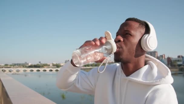 Primer plano del brutal hombre afroamericano bebiendo agua de la botella de deportes después de trotar mirando el cielo azul claro a la luz del día - Imágenes, Vídeo
