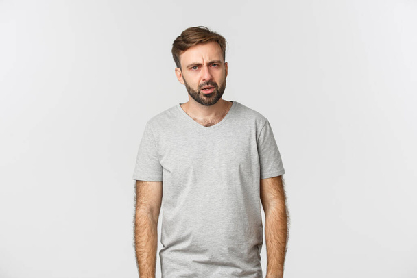 Образ спантеличеного бородатого чоловіка в повсякденній футболці, дивлячись спантеличеним на камеру і хитаючись, не розумієш щось, що стоїть над білим тлом
 - Фото, зображення