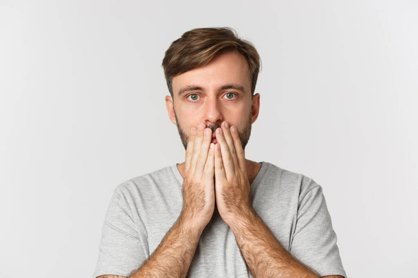 Close-up of shocked and speechless guy, cover mouth and looking at camera, standing over white background - Photo, Image