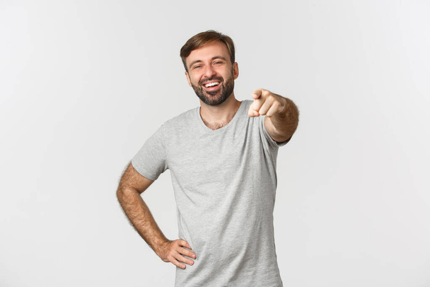 Imagen de un tipo caucásico despreocupado con barba, vistiendo una camiseta gris, riendo y señalando con el dedo a la cámara, de pie sobre fondo blanco - Foto, imagen