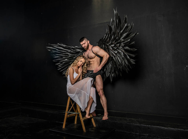 bello uomo muscoloso in costume angelo con ali nere e fragile indifeso bella ragazza in bianco - Foto, immagini