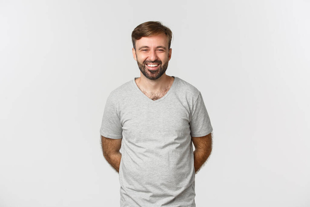 Образ щасливого бородатого чоловіка в сірій футболці, посміхаючись і сміючись, стоїть скромно з руками позаду
 - Фото, зображення