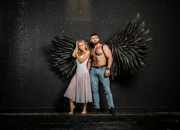 handsome muscular man in angel costume with black wings and fragile defenseless beautiful girl in white - Photo, Image