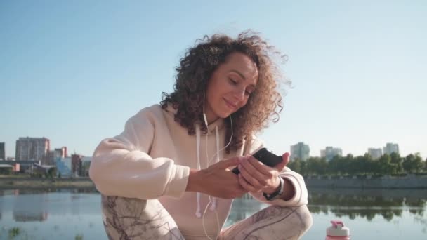 Közepes közelről fiatal női sportoló mosolyog, miközben szörfözés net az ő smartphone ülő és pihentető után szabadtéri kocogás közelében folyó napsütéses reggel - Felvétel, videó