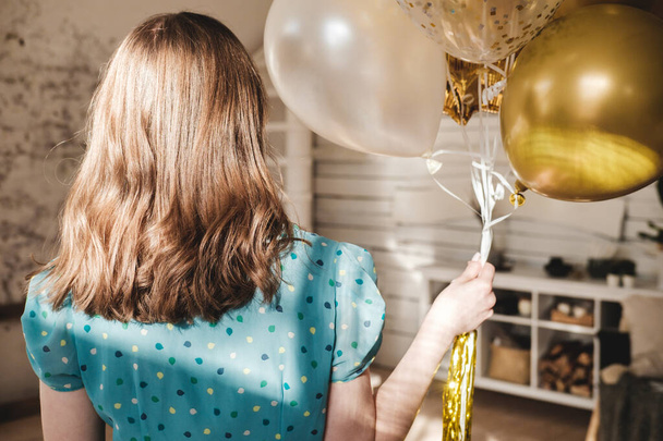 Young beautiful girl in blue dress with white polka dots celebrates her birthday and enjoys the golden balloons. Birthday alone at home during self-isolation. - Φωτογραφία, εικόνα