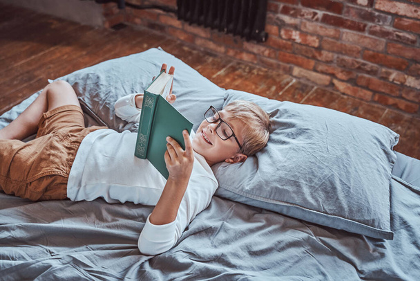 Positiver Junge liest im Wohnzimmer ein Buch und legt sich auf ein Bett - Foto, Bild