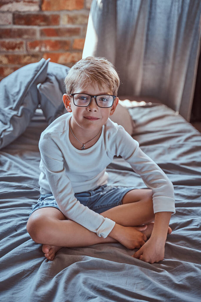 Θετικό αγόρι με γυαλιά κάθεται στο κρεβάτι στο διαμέρισμα - Φωτογραφία, εικόνα