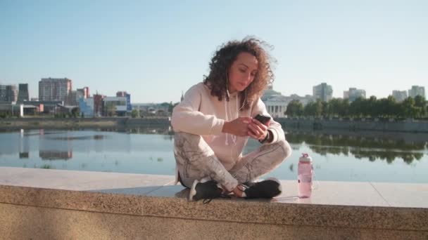 Full shot of cheerful caucasian woman in sportswear checking phone chilling while sitting at embankment drinking water after hard workout and jogging - Footage, Video