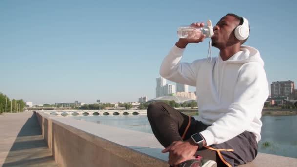 Tiro medio del hombre afroamericano joven que bebe agua de la botella plástica de los deportes que tiene descanso después del entrenamiento al aire libre duro con el horizonte pintoresco en el fondo - Imágenes, Vídeo