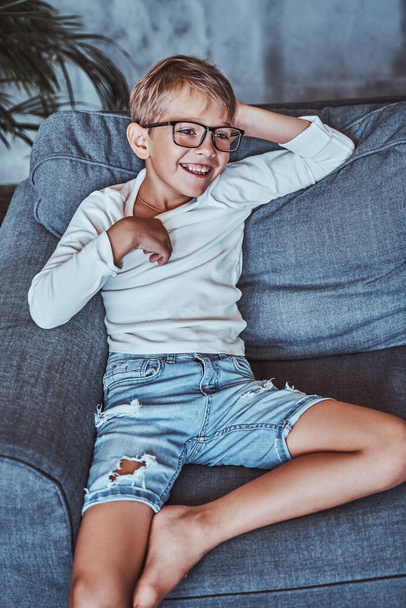 Улыбающийся мальчик в очках позирует на диване в гостиной - Фото, изображение