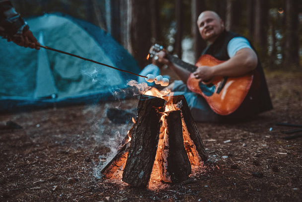 Marshmallow prostet am Lagerfeuer zu und ein Mann mit Glatze spielt Gitarre - Foto, Bild