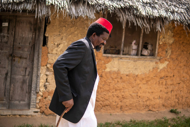 Egy afrikai idősebb férfi a piros muszlim Taqiyyah Fez kalap és blézer Sétál egy bottal bicegő emberek közelében az alapvető kunyhó Thatched tető Kis távoli falu Tanzánia, Pemba sziget, Zanzibar - Fotó, kép