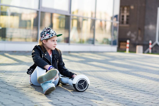 Young thoughtful girl in a baseball cap sitting on the sidewalk with a hoverboard. - Foto, afbeelding