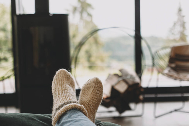 Feet in warm knitted woolen socks on background of modern black fireplace and big windows with view on mountains. Woman relaxing in comfortable home, cozy warm moments - Foto, immagini