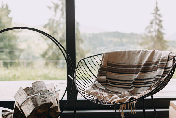 Moderne zwarte stoel met gezellige deken en brandhout op metalen statief op de achtergrond van grote ramen met uitzicht op de bergen. Gezellige warme en rustige momenten in de cabine tijdens het koude seizoen. - Foto, afbeelding