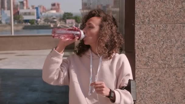 Medium shot of tired exhausted woman after hard workout sipping water from special sports bottle leaning on outdoor column hiding from sun - Footage, Video