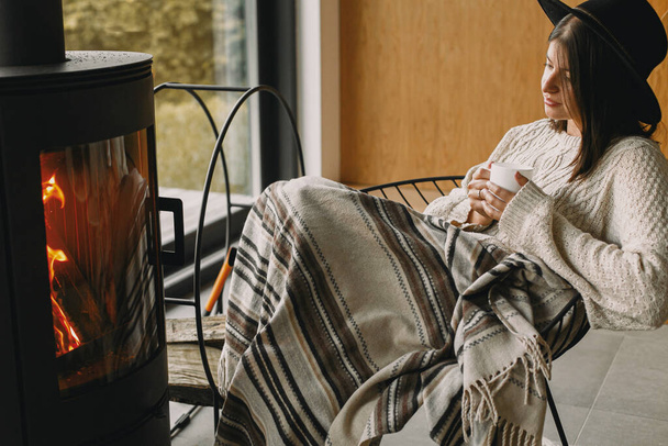 Стильна жінка в в'язаному светрі і капелюсі тримає теплу чашку чаю в сучасному чорному каміні з видом на гори. Затишні теплі моменти в холодну пору року. Молода хіпстерка розслабляється в комфортному будинку
 - Фото, зображення
