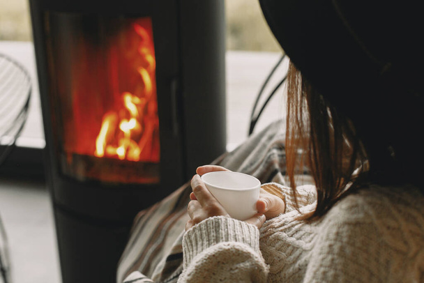Mujer elegante sosteniendo una taza de té caliente y relajante en la moderna chimenea negra con vista a las montañas. Acogedores momentos cálidos en temporada fría. Mujer joven en jersey de punto y sombrero descansando - Foto, imagen