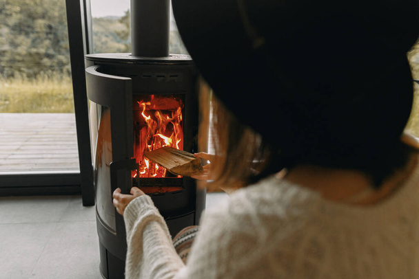 Femme élégante en pull tricoté mettant du bois de chauffage à cheminée noire moderne avec feu chaud. Moments chauds et confortables à la saison froide. Hipster femelle relaxant dans une cabine confortable en montagne - Photo, image