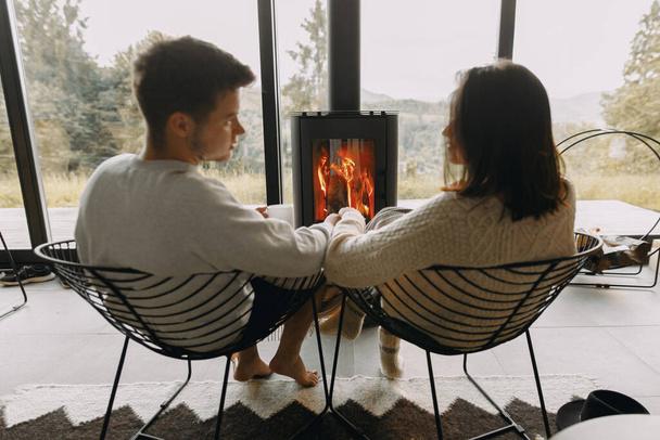 Couple élégant en chandails avec des tasses de thé chaud relaxant à la cheminée noire moderne avec vue sur les montagnes. Des moments chaleureux et chaleureux en famille à la saison froide. Jeune famille en vacances - Photo, image