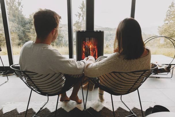 Stilvolles Paar in Pullovern entspannt zusammen am modernen schwarzen Kamin mit Blick auf die Berge. Gemütliche warme Familienmomente in der kalten Jahreszeit. Junge Familie im Urlaub - Foto, Bild
