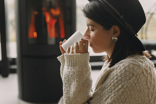 Stylish woman in knitted sweater drinking warm cup of tea and relaxing at modern black fireplace with view on mountains. Cozy warm moments at cold season. Young female enjoying drink - Photo, Image