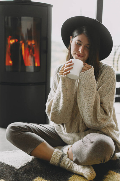 Κομψή γυναίκα με πλεκτό πουλόβερ και καπέλο που κρατά ζεστό φλιτζάνι τσάι και χαλαρώνει στο μοντέρνο μαύρο τζάκι με θέα στα βουνά. Ζεστές στιγμές στην κρύα εποχή. Νεαρή γυναίκα ξεκουράζεται - Φωτογραφία, εικόνα