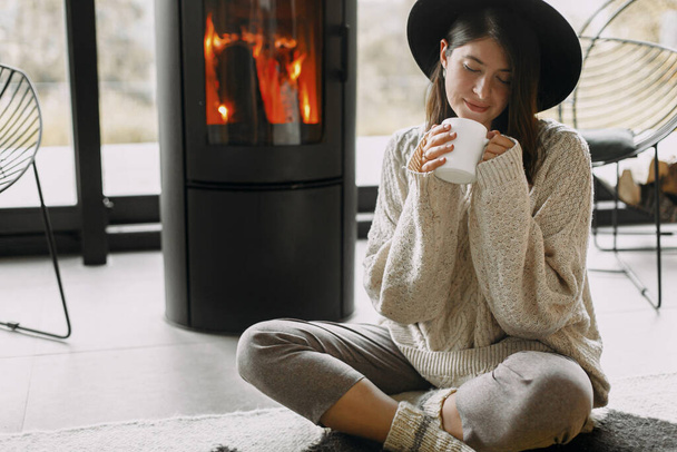 Κομψό γυναίκα κρατώντας ζεστό φλιτζάνι τσάι και χαλαρώνοντας στο σύγχρονο μαύρο τζάκι με θέα στα βουνά. Ζεστές στιγμές στην κρύα εποχή. Νεαρή γυναίκα με πλεκτό πουλόβερ και καπέλο αναπαύεται - Φωτογραφία, εικόνα