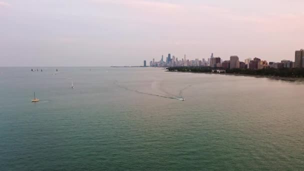 Vista aérea panorámica de una lancha a motor que cruza un tranquilo lago Michigan mientras los veleros flotan antes del atardecer con el horizonte de Chicago y los edificios de gran altura en el horizonte más allá. - Imágenes, Vídeo