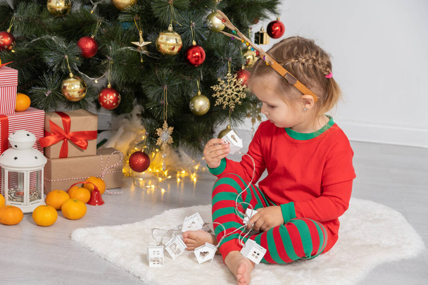 toddler girl with headband with horns of a deer sitting near the Christmas tree. - 写真・画像