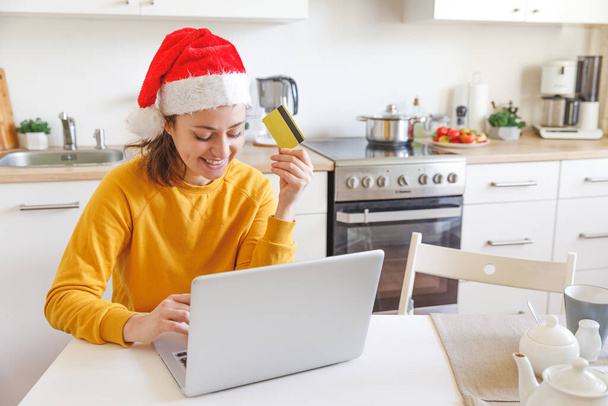Woman in Santa hat shopping online and paying with gold credit card. Young girl with laptop buying Christmas gift present on Internet on kitchen indoor. Online Christmas shopping concept - Photo, Image