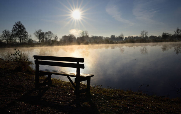 日の出には湖のそばに古い木のベンチが立ち、秋には霧が立ち上がります。 - 写真・画像