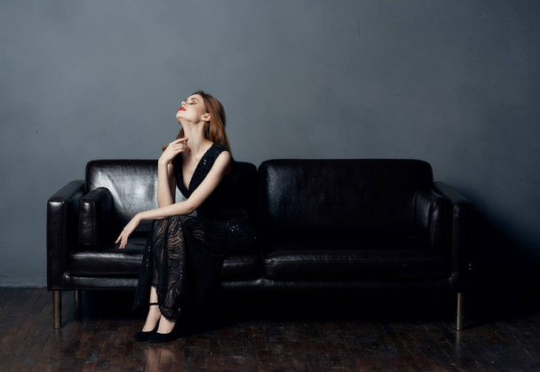 Kobieta w czarnej sukience siedzi na skórzanej kanapie w domu makijaż wieczorem nagie ramiona model - Zdjęcie, obraz