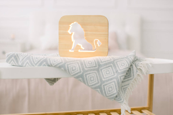 Front close up view of stylish wooden night lamp with lion cut out picture, on gray blanket at cozy light bedroom interior. Wooden decorations at home interior - Φωτογραφία, εικόνα