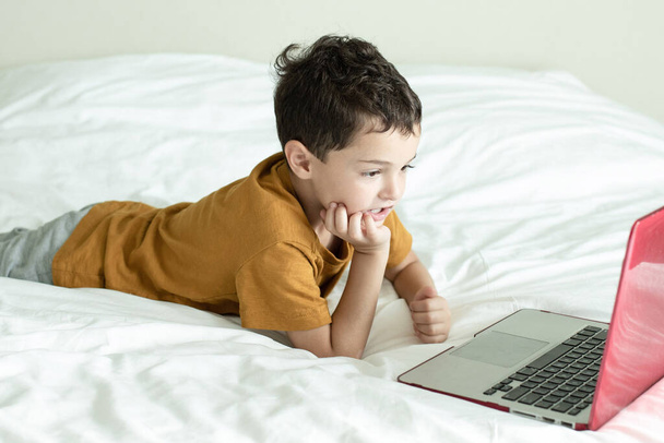 Le concept d'apprentissage et de jeu des enfants sur l'ordinateur, les enfants et les écrans, l'éducation à la maison. Un enfant européen couché sur un lit blanc regarde l'écran de l'ordinateur portable. Horizontal. - Photo, image