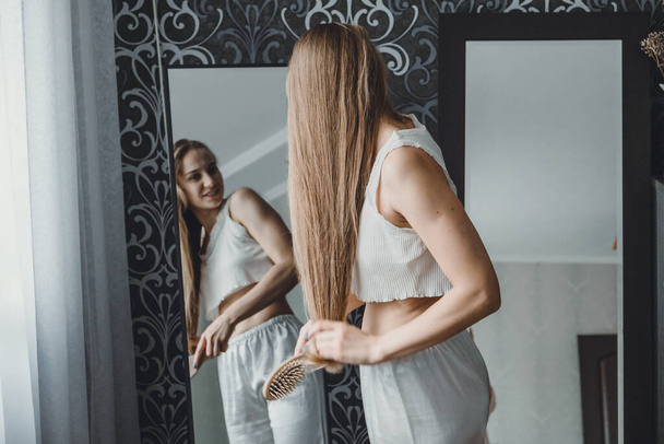 Cómo cuidar el cabello largo. Vista posterior de la joven rubia natural en ropa de casa blanca peinándose el pelo y mirando al espejo en el dormitorio - Foto, imagen