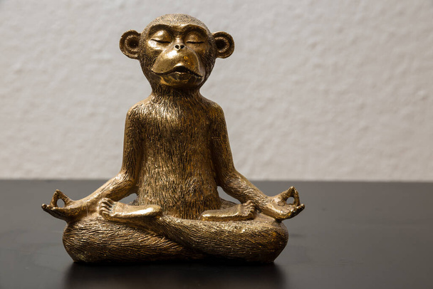 A gold colored meditating monkey statue sitting on a black surface, the background of the picture is white and blurry. - Photo, Image