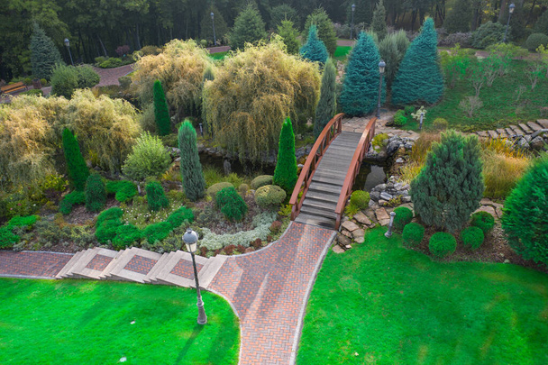 Landscape design of a public park. Wooden bridge across a dry river. Various ornamental plants grow on rockeries. Green lawn near the paved paths. Great place to relax. - Photo, Image
