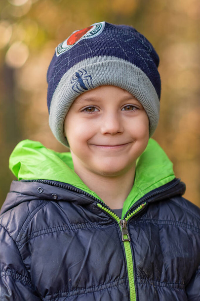 Šťastný chlapeček s kloboukem a zimní bundou pózuje o podzimním dni. Fotografie v lese s krásným bokeh. - Fotografie, Obrázek
