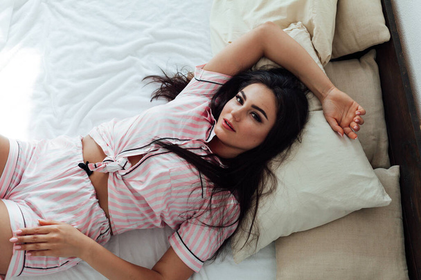 Beautiful brunette woman in pink pajamas on bed in bedroom after sleeping - Fotoğraf, Görsel