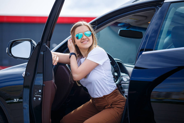 長い髪の若い美しい女性は、駐車場で黒い車の中に座っています。カジュアルな服を着た可愛い女の子。レンタカー旅行 - 写真・画像