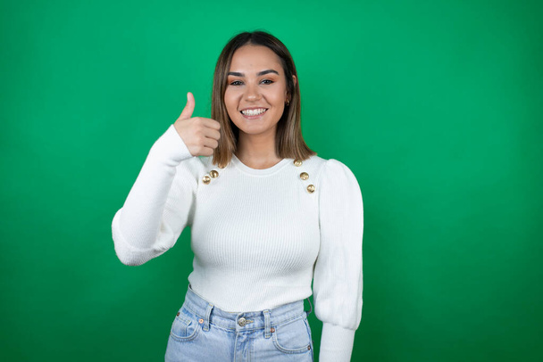 Young beautiful woman wearing white sweater over isolated green background smiling and doing the ok signal with her thumb - Photo, Image
