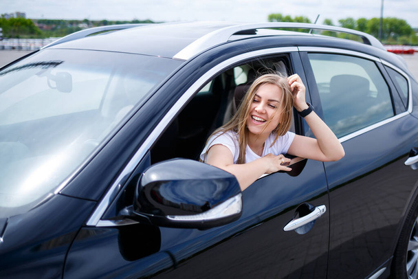 Young beautiful woman with long hair sits in a black car at a parking lot. Pretty girl in casual clothes. Car trip - Photo, Image