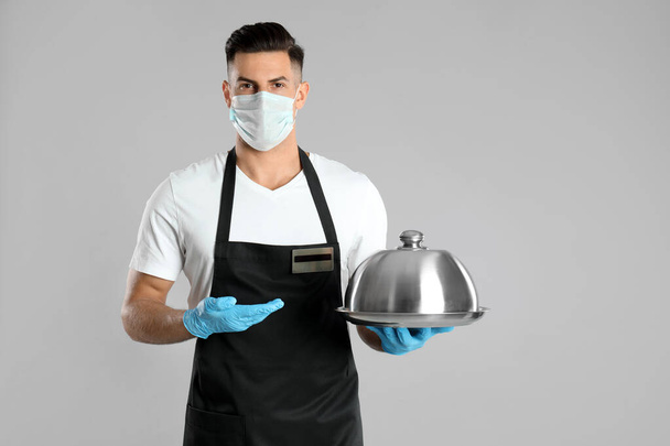 Waiter in medical face mask holding tray with lid on light grey background - Fotoğraf, Görsel