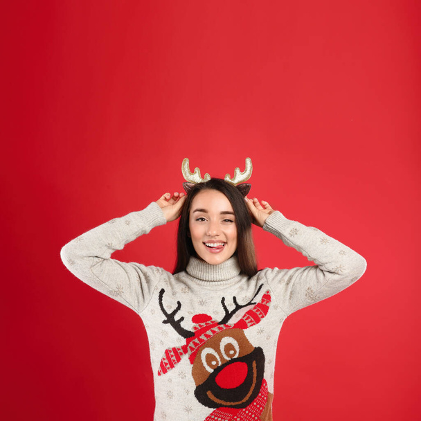 Young woman in Christmas sweater and deer headband on red background, space for text - Фото, зображення